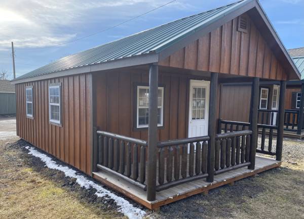 Small wood cabin with brown trim and log railing, covered front porch, and white 9 lite door, set on gravel pad at our Blakely sales lot.